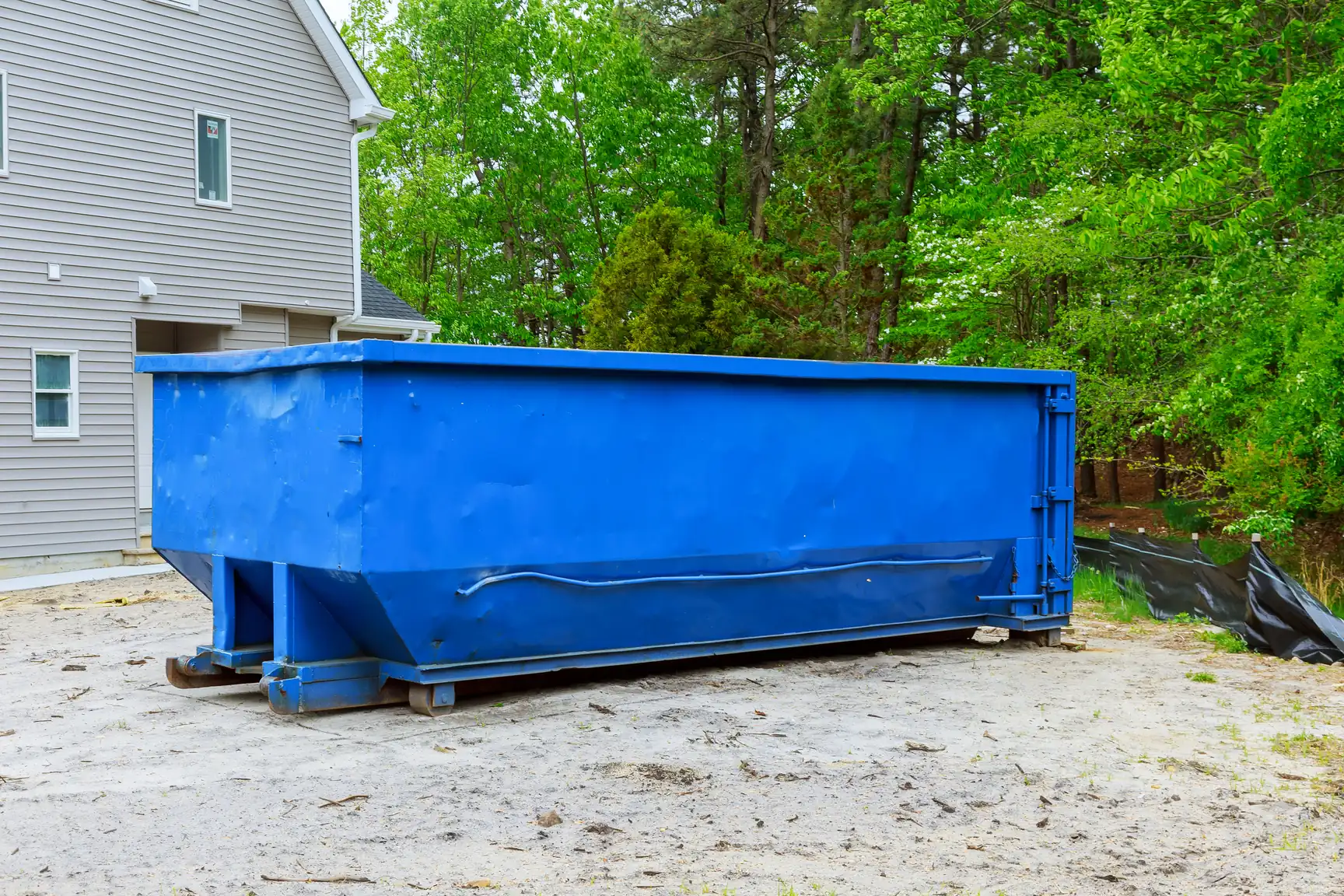 An empty blue roll-off container on sand beside a house under construction